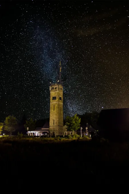 Illuminated stone tower at Signal de Botrange beneath a star-filled sky, with the Milky Way above and a dark foreground.