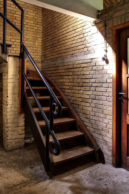 Narrow wooden stairs against a brick wall, with a black handrail and a lamp above.