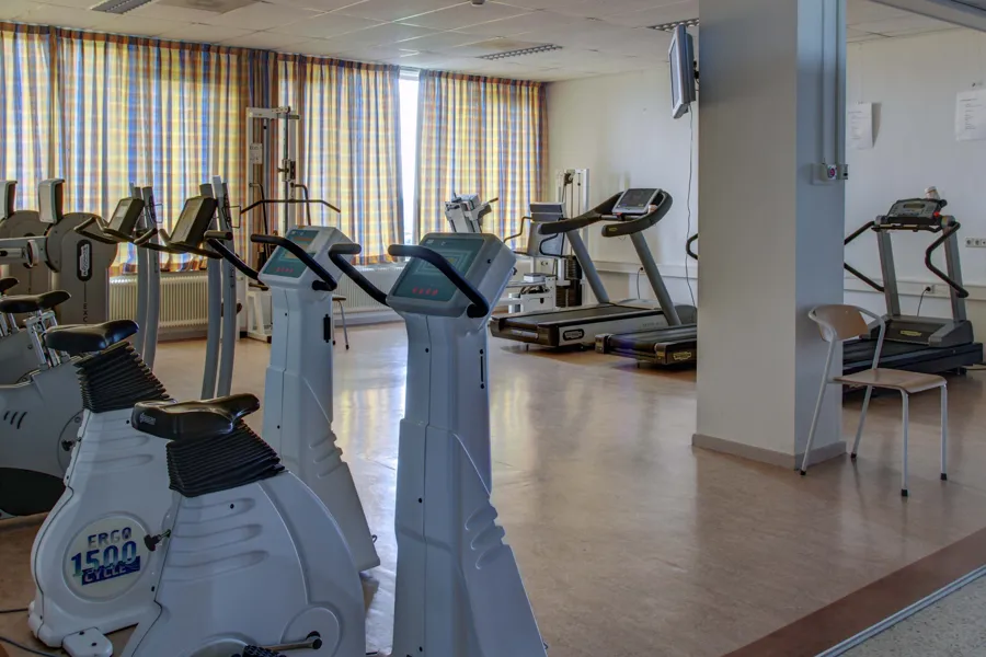 Ergometer und Laufbänder in hellem Rehabilitationsraum mit Holzboden und großen Fenstern mit Vorhängen.