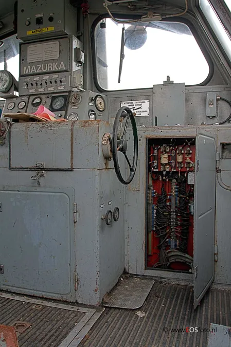 Grijze locomotiefcabine met stuurwiel, meters en een open elektrische kast.