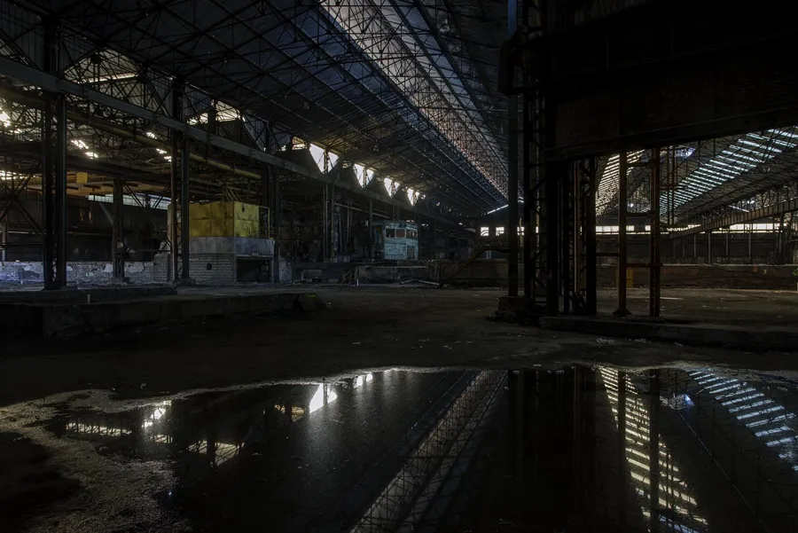 Flaque reflétant le toit d’une halle industrielle sombre.