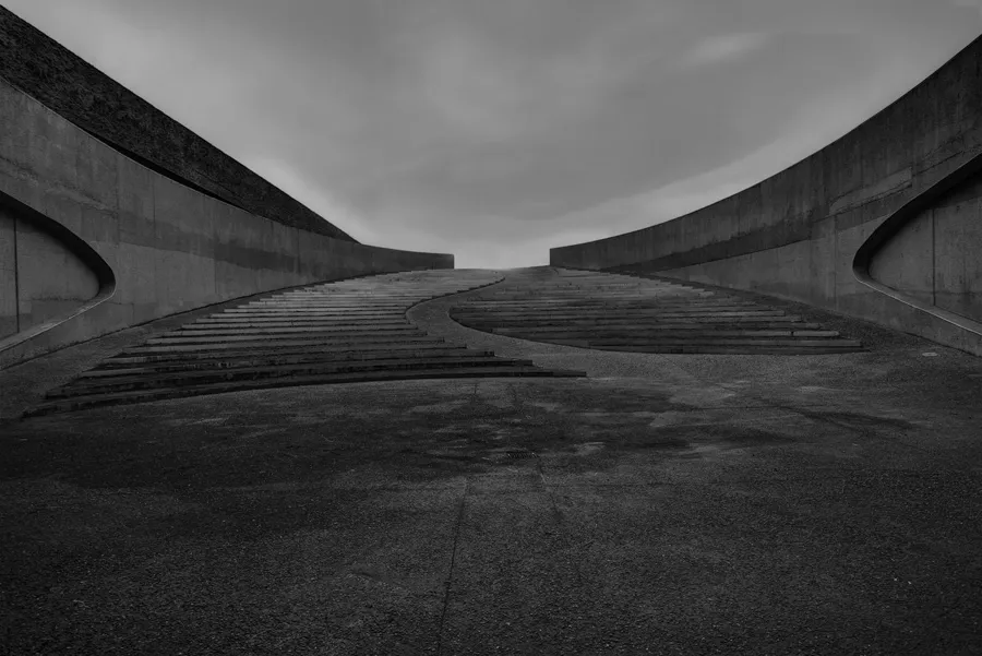 Zwart-witbeeld vanaf de voet van een brede trap, met een slingerende middenstrook en naar buiten gebogen betonwanden.