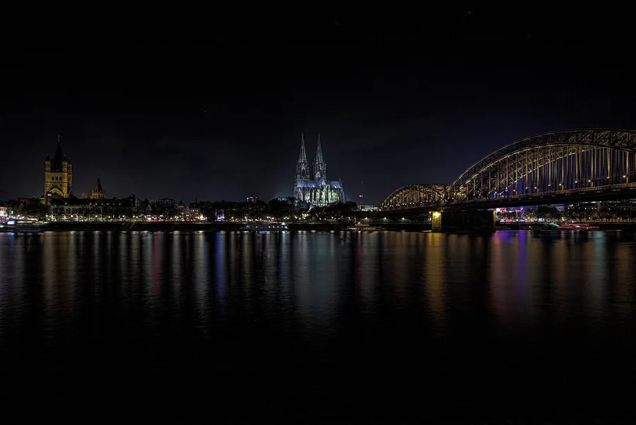 Wide night view of Cologne’s Rhine waterfront, with a lit tower on the left, the cathedral in the centre and the Hohenzollern Bridge on the right above dark water.