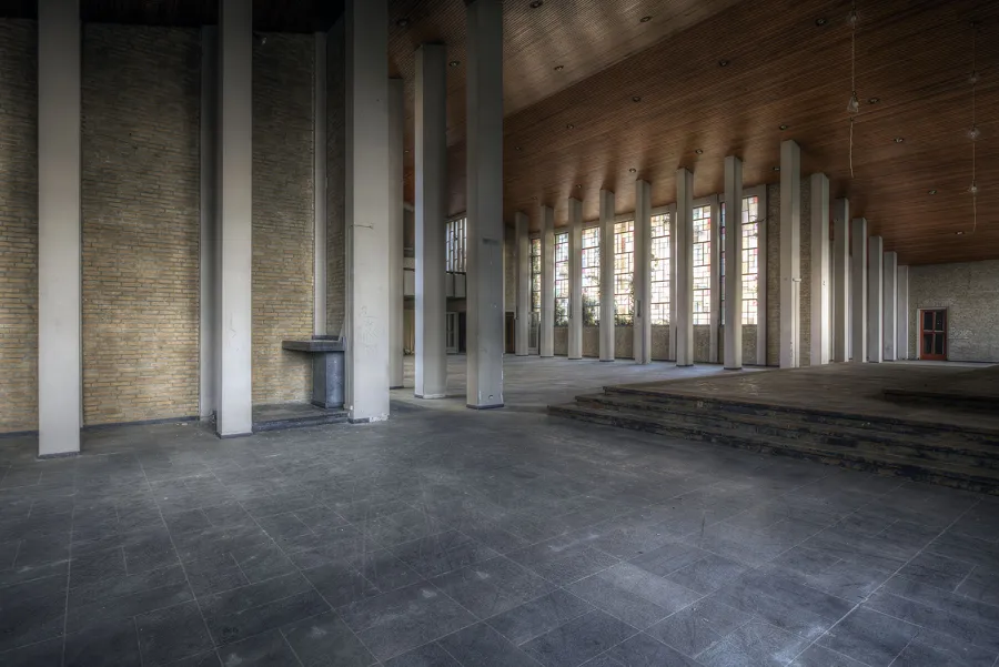 Sala amplia con tejado de madera a dos aguas, columnas y plataforma al fondo.