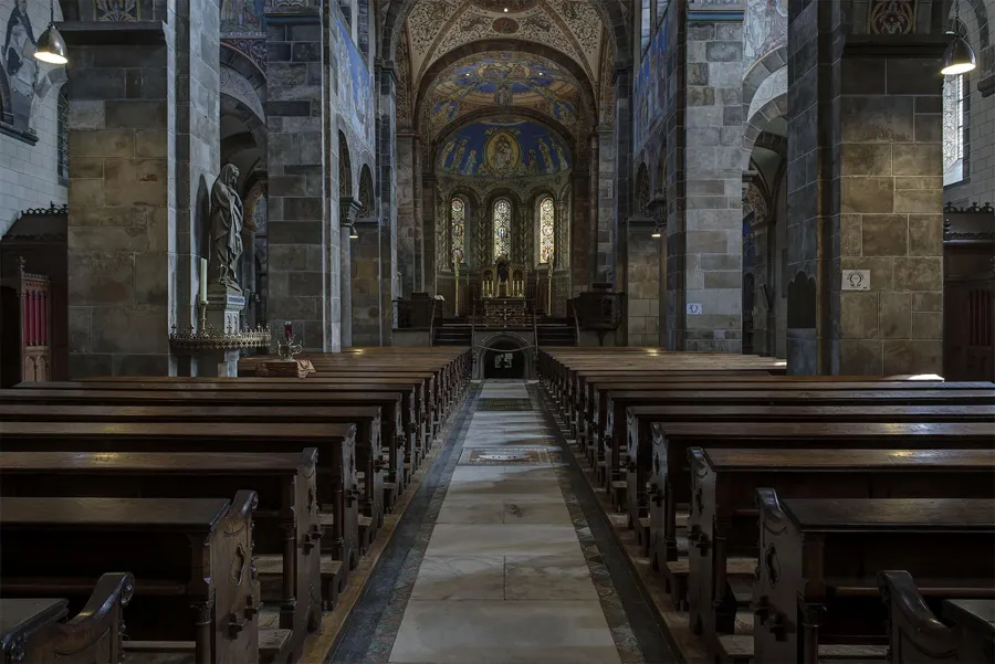Mittelgang der Abteikirche Rolduc mit Holzbänken zwischen mächtigen Steinsäulen unter einem bemalten Gewölbe.