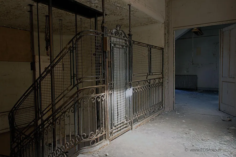 Grille métallique ouvragée autour d’un ancien ascenseur près d’une porte ouverte.
