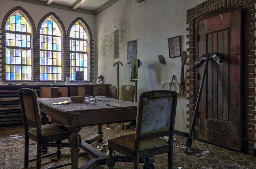 Sakristeiraum mit Holztisch, Stühlen, hohen Fenstern und alter Tür.