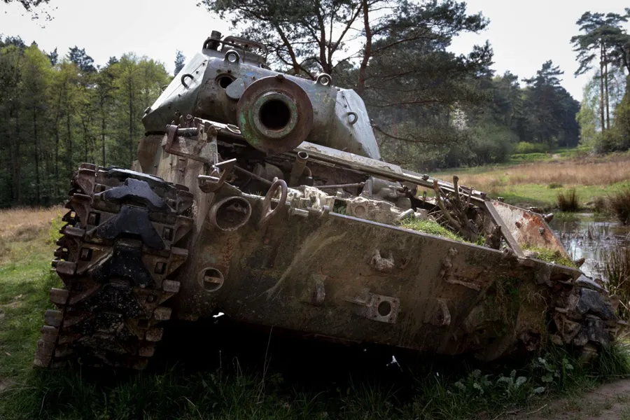 Beschädigter Panzer mit rostigen Ketten im Gras am Waldrand.