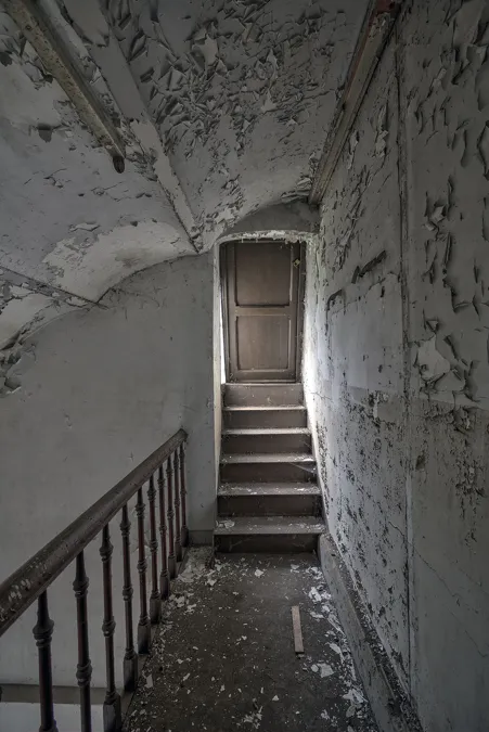 Escalera estrecha con paredes desconchadas hacia una puerta de madera.