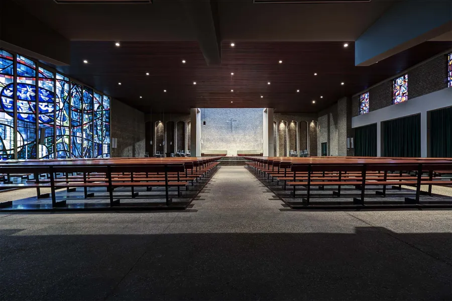 Wide church interior with a glass wall left, pews and a pale wall behind the altar area.