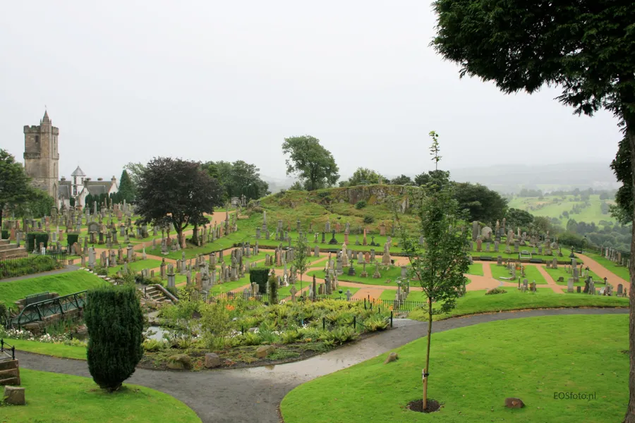 Grüner Friedhof am Hang mit Wegen, Grabsteinen und Bäumen sowie einem Steinturm links oben.