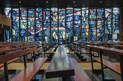 Large blue, red and green stained-glass wall behind long wooden pews.