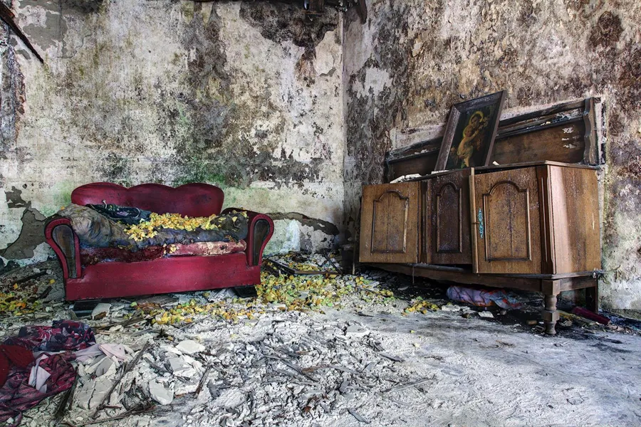 Red sofa and wooden cabinet in a room with peeling walls and rubble on the floor