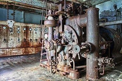 Machine industrielle rouillée avec de hauts cylindres, une grande roue et de nombreux tuyaux dans une halle délabrée.