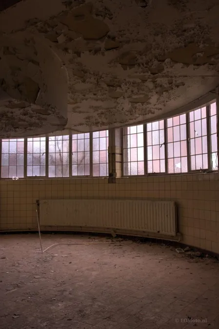 Geschwungene Wand mit hohen rosa Fenstern, Heizkörper und abblätternder Decke.