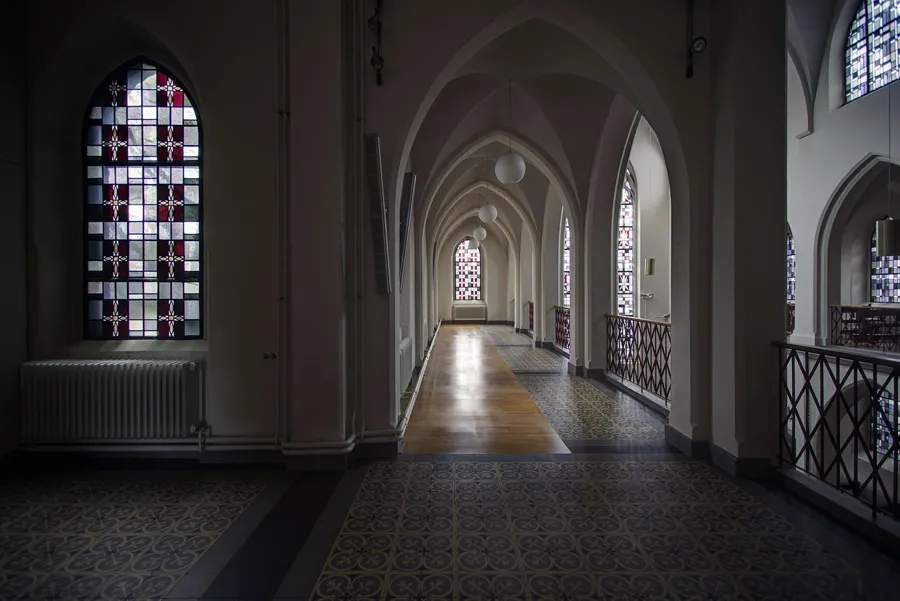 Lange galerij met spitsbogen, kleurrijk raam links en metalen balustrade rechts.