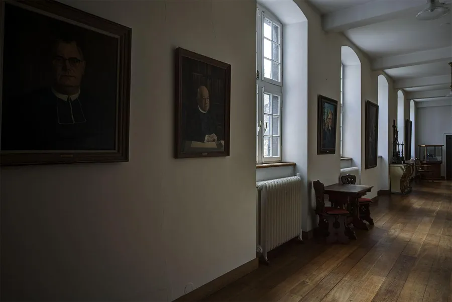 Dunkle Porträts in einem Klosterflur mit hohen Fenstern, Heizkörpern und glänzendem Holzboden.