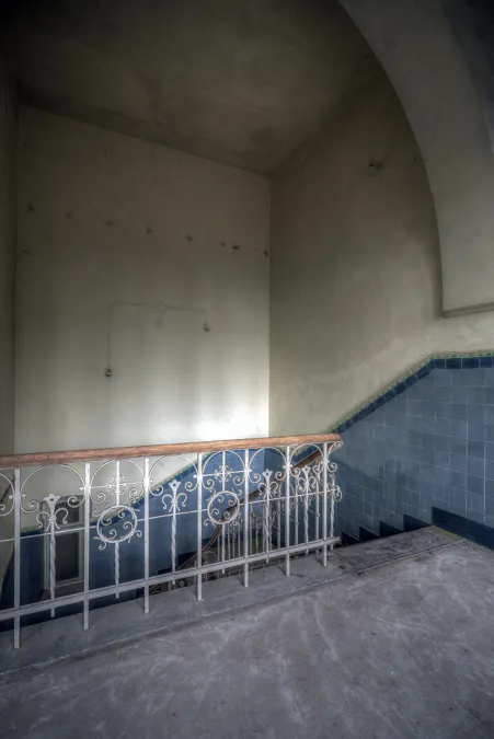 Escalera estrecha descendente con paredes inferiores azules y barandilla decorada.