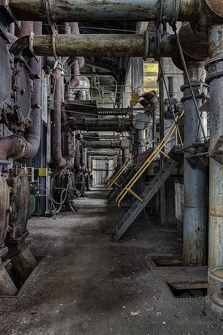 Narrow passage between dense pipework and stairs with a yellow railing.