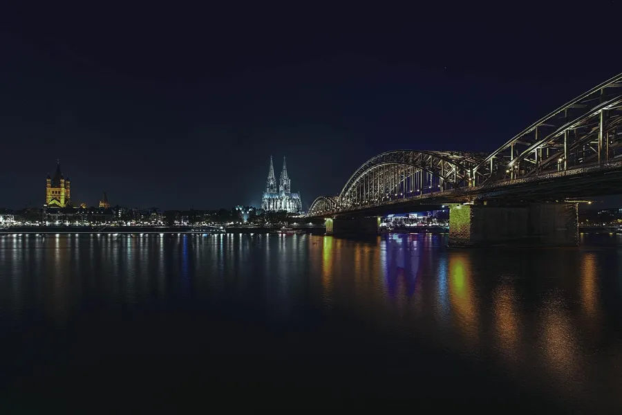 Night photograph along the Hohenzollern Bridge leading from the right towards the illuminated cathedral, with coloured streaks of light on the smooth-looking Rhine.