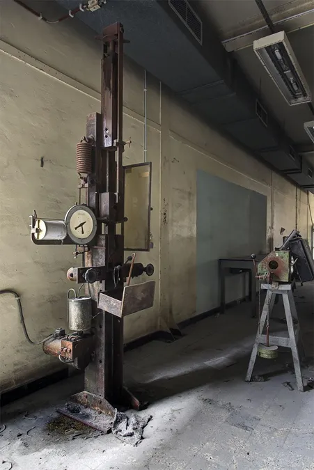 Hoge testmachine met ronde meter bij een schoolbord.