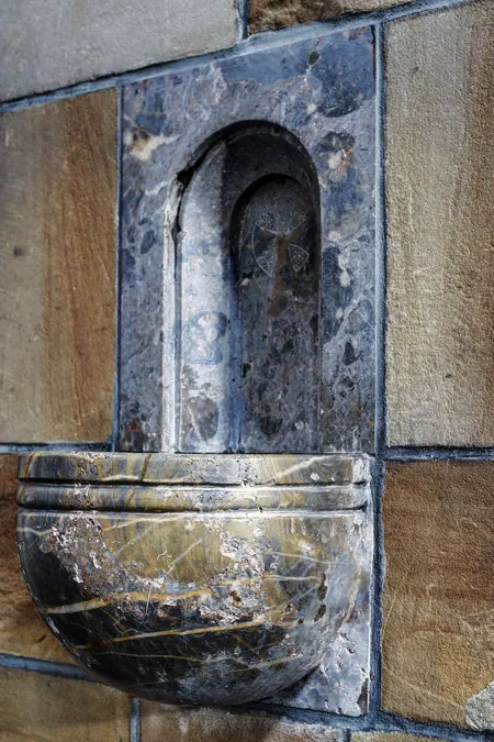 Pequeña pila de agua bendita de piedra en un muro, bajo un nicho oscuro con cabeza esculpida.