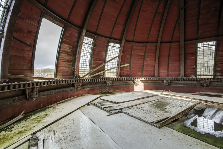 Sala circular con cúpula de madera roja, ventanas altas y suelo deteriorado.
