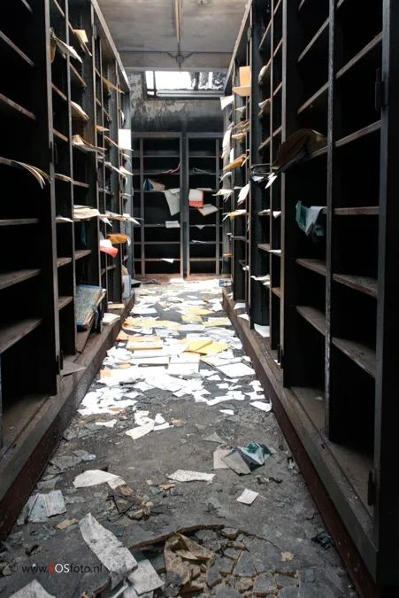 Allée d’archives étroite entre de hautes étagères aux papiers épars.