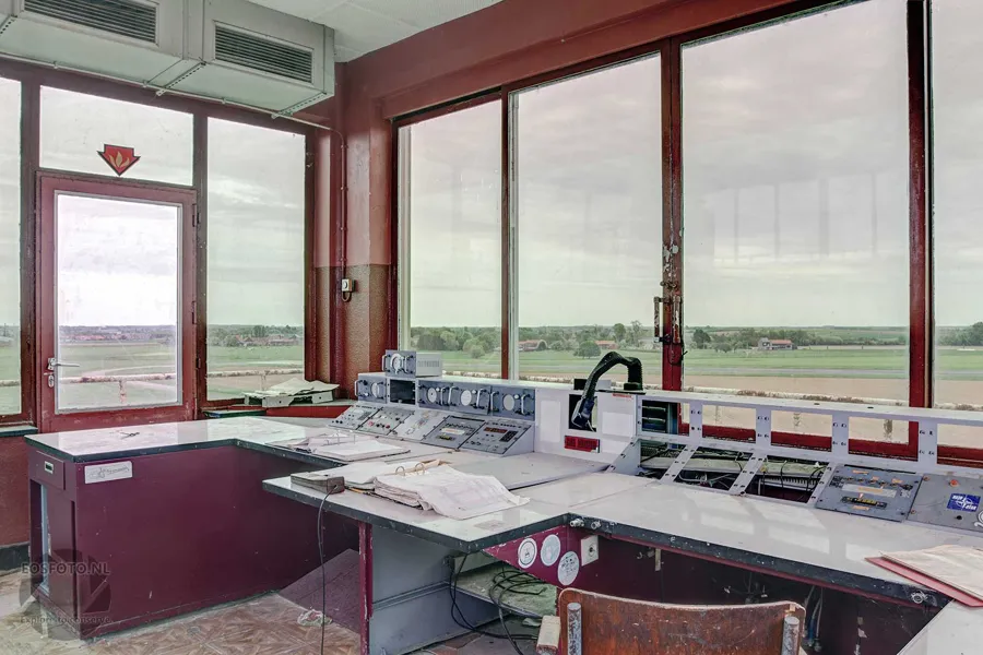 Broad control desk with gauges and switches beside red window frames.