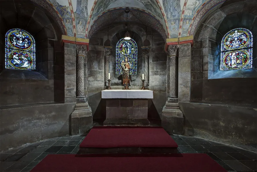 Niedriger Gewölberaum mit weißem Altar zwischen Steinsäulen und drei farbigen Glasfenstern.