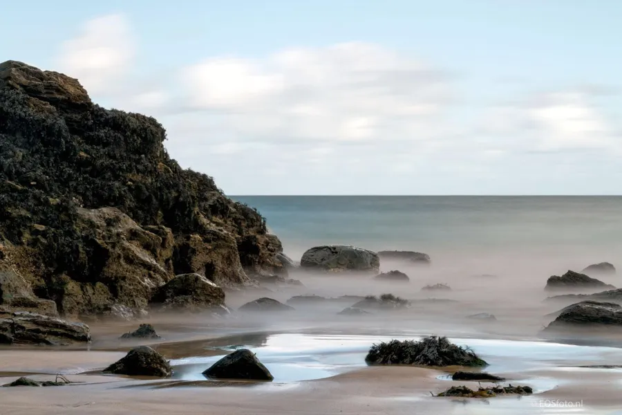 Dunkle Küstenfelsen an einem kleinen Strand, mit durch Langzeitbelichtung geglättetem Meerwasser dazwischen.