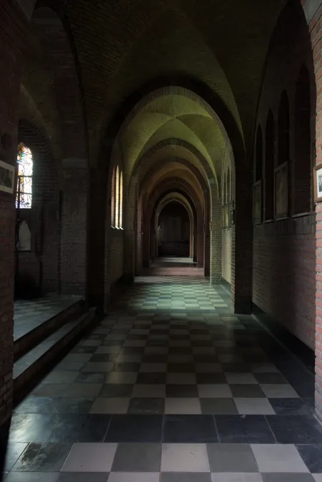 Pasillo monástico oscuro con arcos sucesivos y suelo ajedrezado.