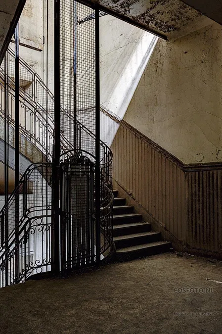 Schmaler alter Aufzugskäfig neben einer Treppe im Tageslicht.