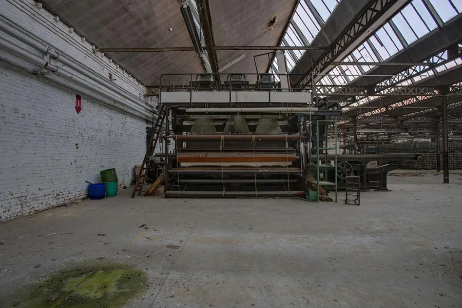 Máquina textil bajo un tejado en diente de sierra junto a una zona vacía.