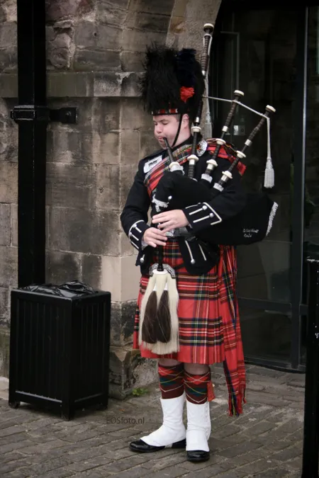 Dudelsackspieler mit Kilt, hoher schwarzer Mütze und weißen Kniestrümpfen an einem Steineingang.
