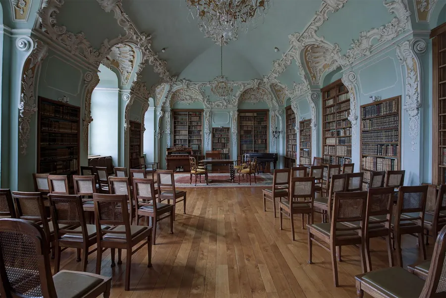 Hellblauer Bibliothekssaal mit weißem Stuck, hohen Bücherschränken und Holzstuhlreihen auf Parkett.
