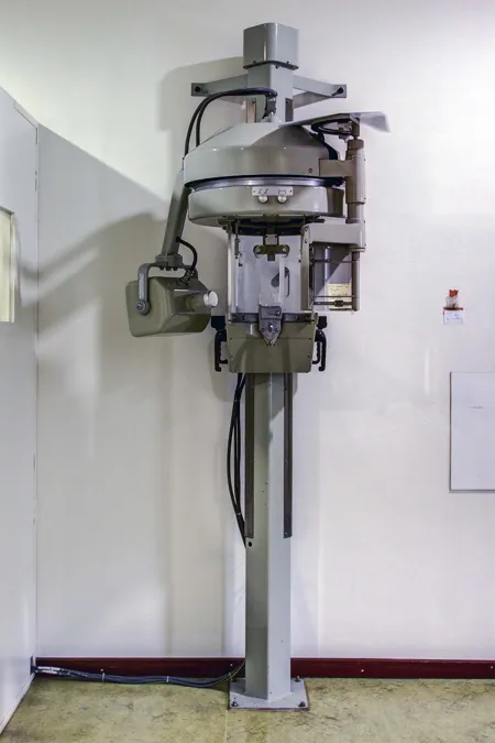 Altes senkrechtes Röntgengerät mit Metallgehäuse und herabhängenden Kabeln vor weißer Wand.