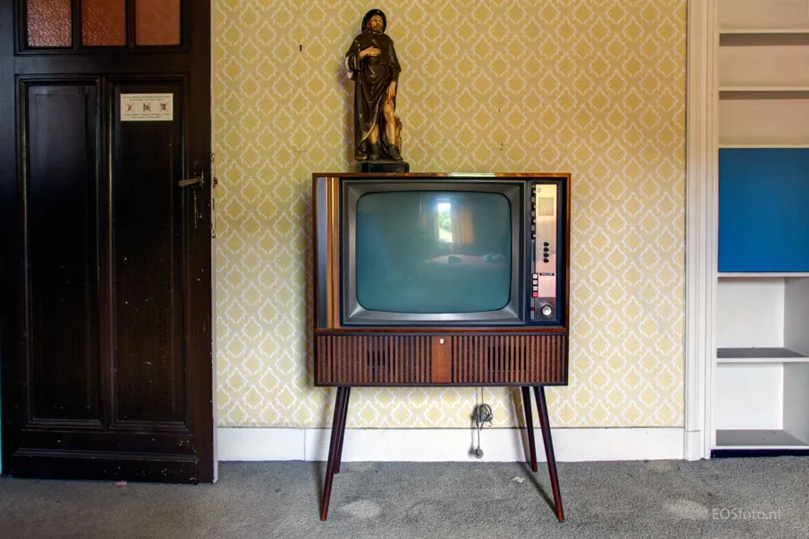 Old television on tall legs with a saint’s statue above it.