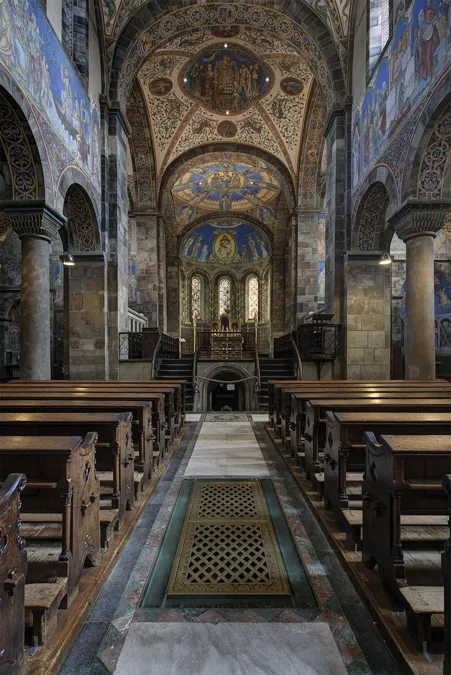 Hochformat durch die Abteikirche mit Bodengittern im Mittelgang und bemalten Bögen über den Bänken.