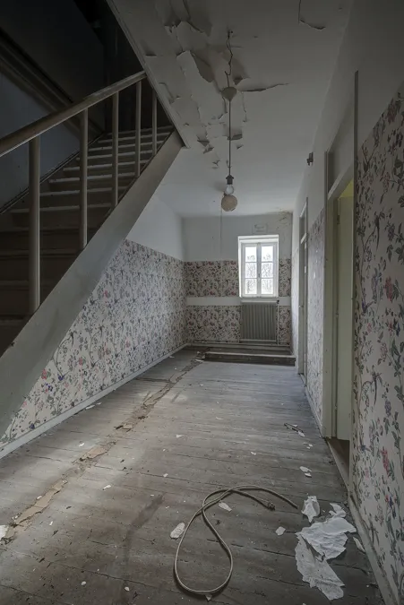 Pasillo deteriorado bajo una escalera con ventana al fondo.