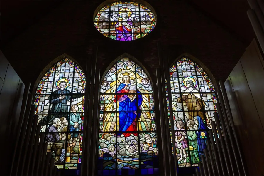 Three stained-glass windows with religious figures beneath a smaller round window in a dark church wall.