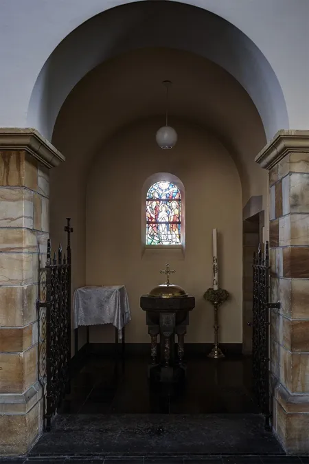 Pila bautismal en una pequeña capilla abovedada, con vidriera estrecha y candelabros detrás.