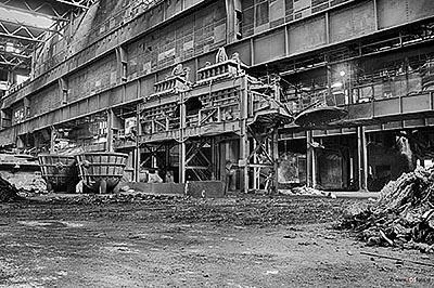 Vista en blanco y negro de una nave industrial abandonada con vigas de acero, tuberías y escombros en el suelo.