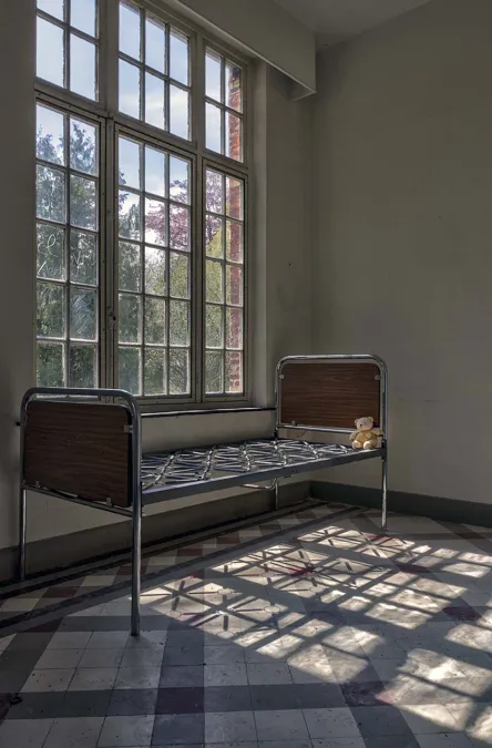 Lit métallique près de hautes fenêtres projetant des ombres sur le sol carrelé.
