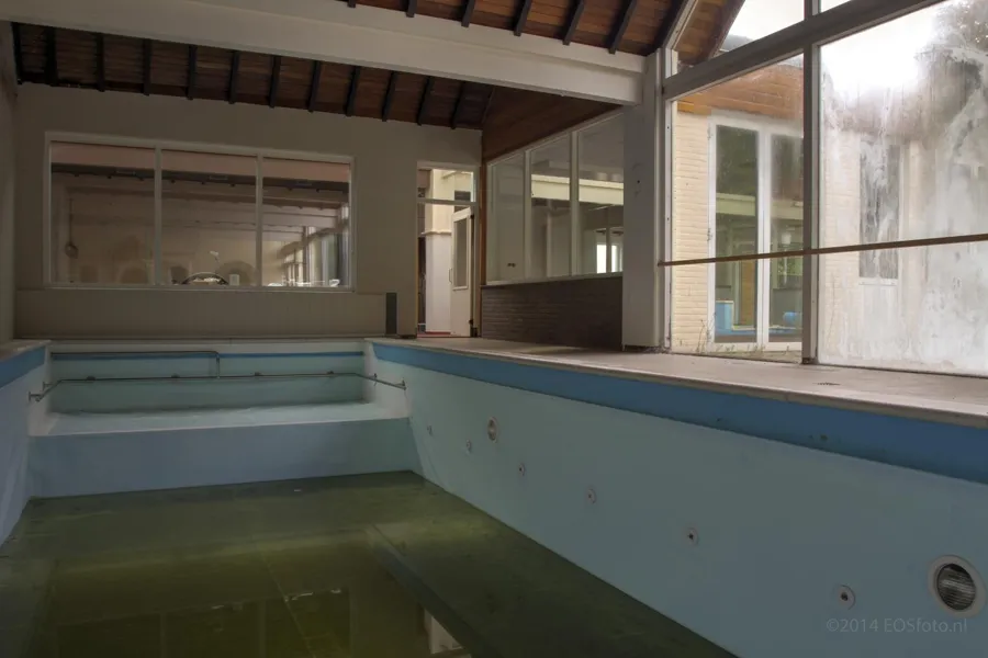 Piscina interior vacía con agua verde en el fondo junto a ventanas altas.