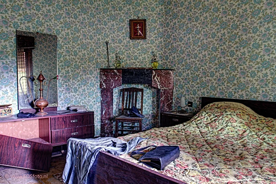 Bed with a floral blanket opposite a marble fireplace and blue floral wallpaper