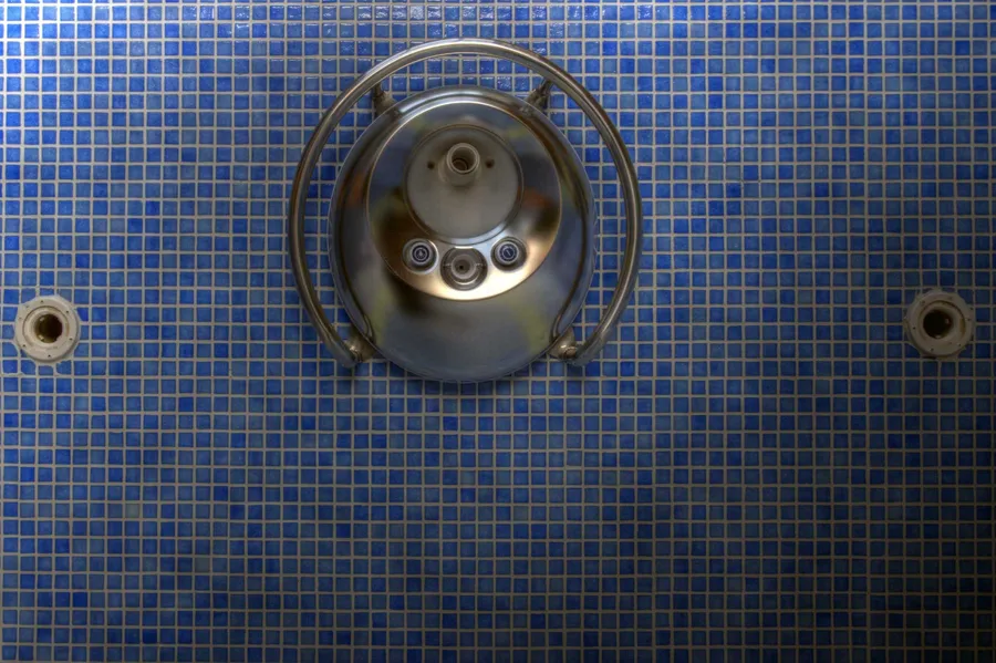 Runder Metallbeckenanschluss mit kleinen Düsen auf blauen Mosaikfliesen.