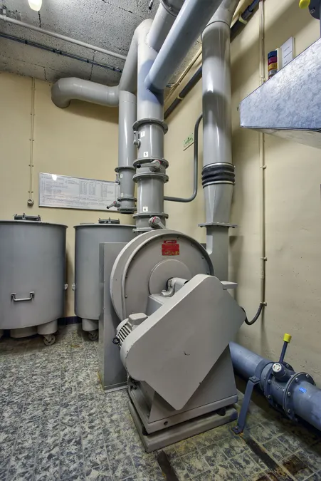 Grijze ventilator met motor en dikke luchtleidingen in een technische ruimte.