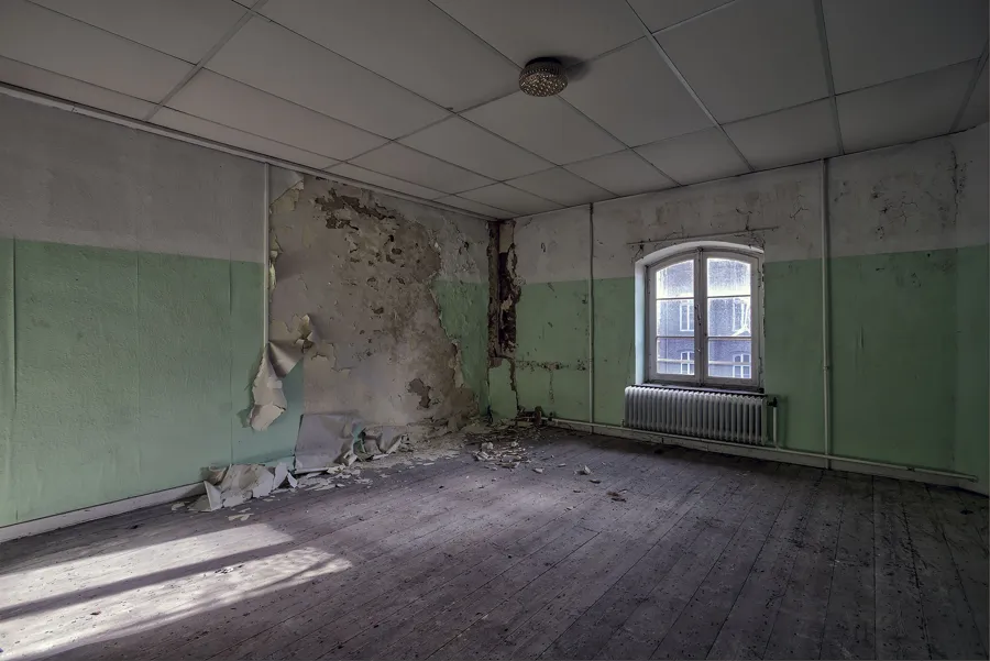 Sala vacía con paredes inferiores verdes, una ventana y yeso desmoronado.