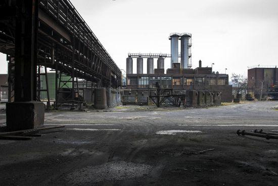 Open factory yard beside a pipe bridge with tall chimneys behind